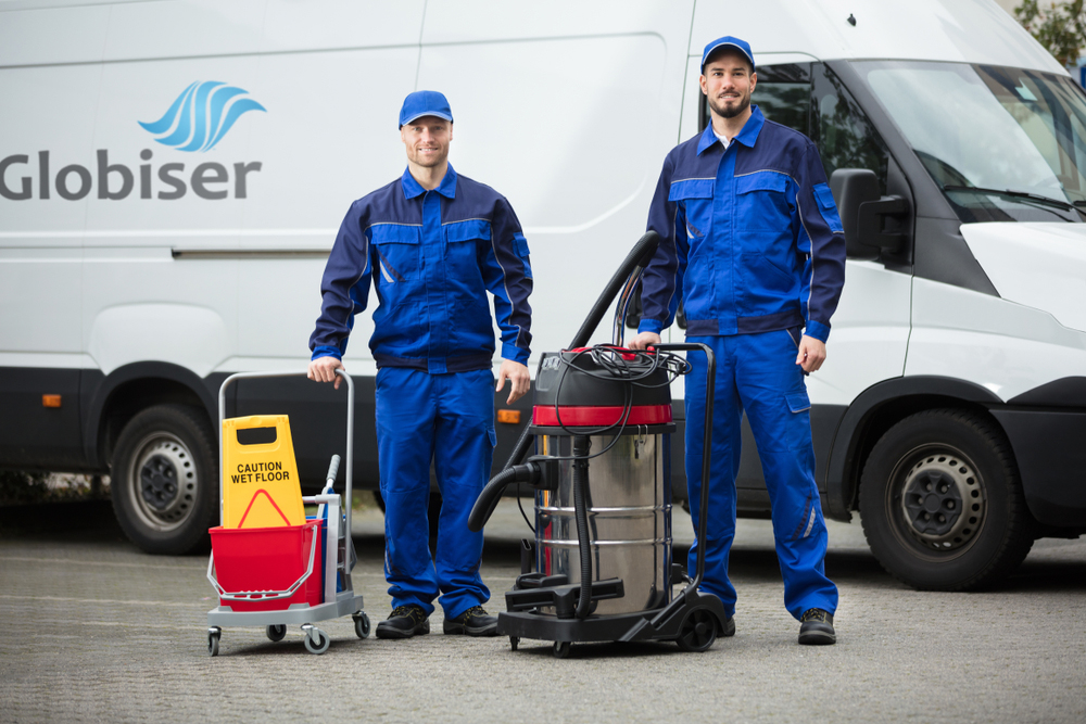 Two happy male Globiser team janitors standing with cleaning equipment in front of a Globiser Van